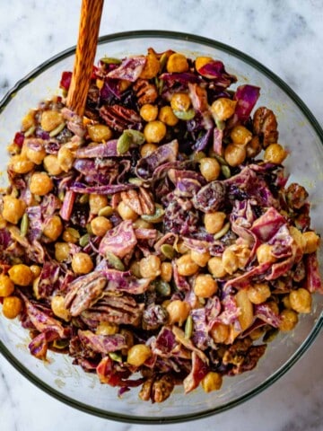 Roasted red cabbage, chickpeas, pecans, pepitas, and dried cranberries in creamy dressing in a glass bowl with a wooden spoon.