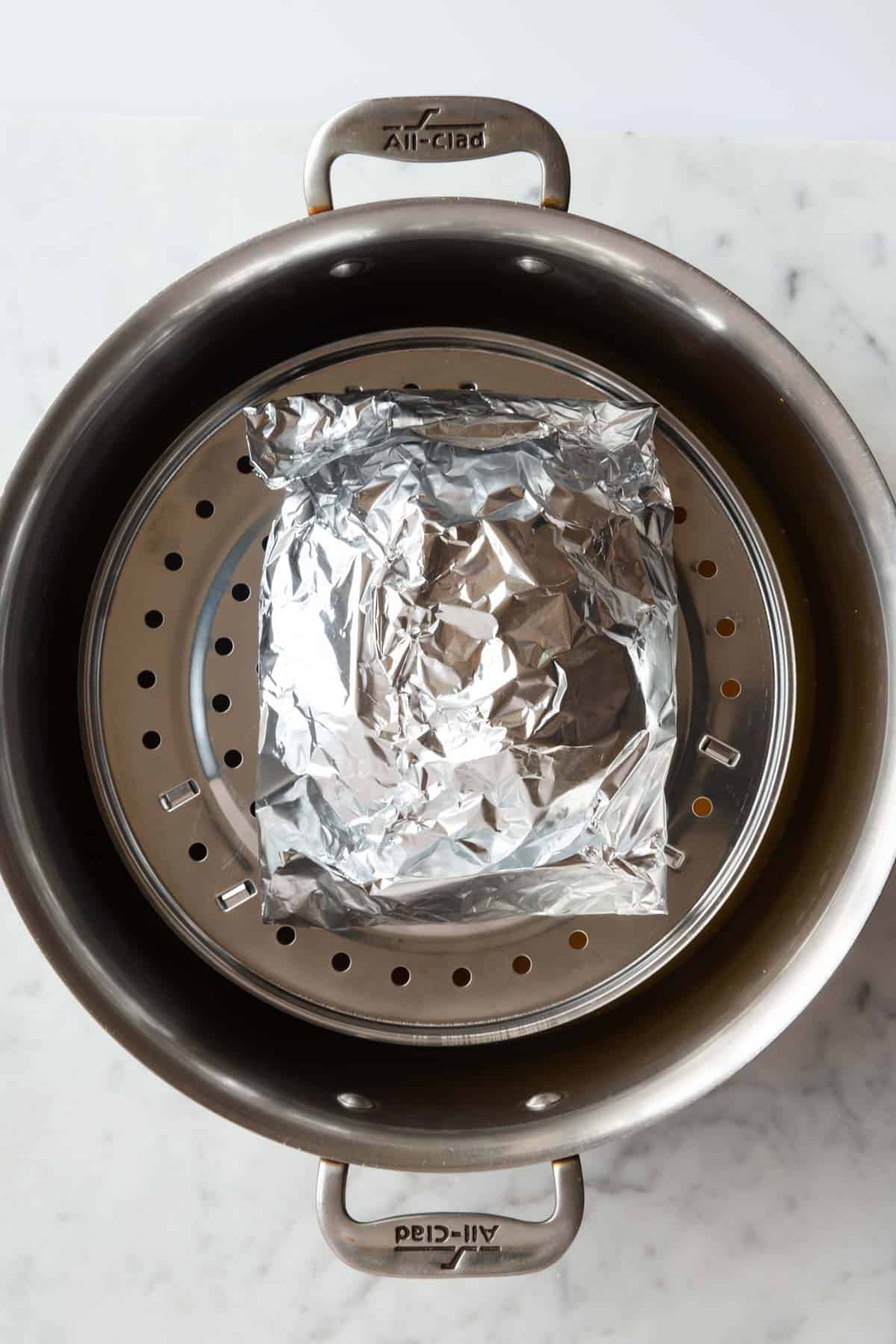 Seitan in aluminum foil being placed on steaming basket in a stainless steel saucepan.