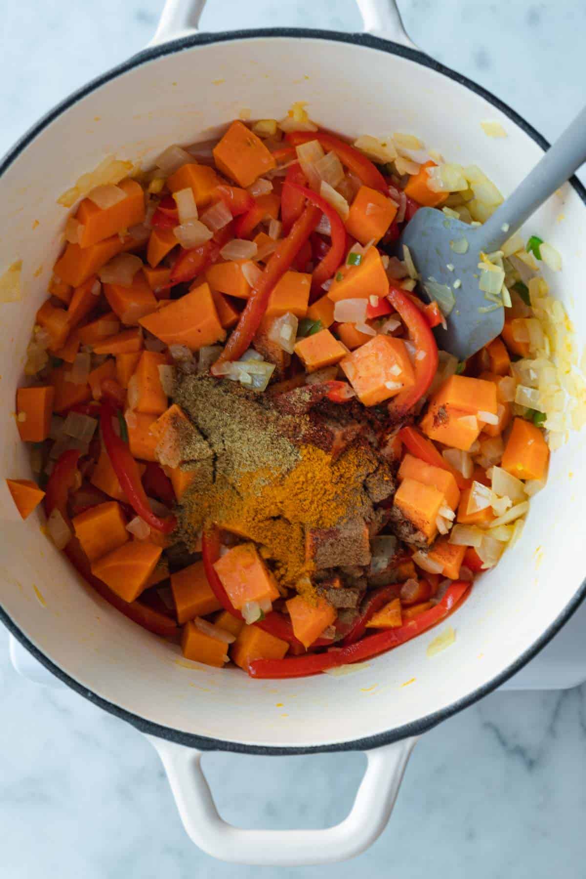 Chopped sweet potatoes, onion, red bell pepper, garlic, ginger, green chili pepper, and spices cooking in a white saucepan with a gray spatula.