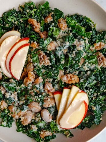 Kale quinoa salad with butter beans and walnuts in a white bowl with creamy dressing and sliced apple.