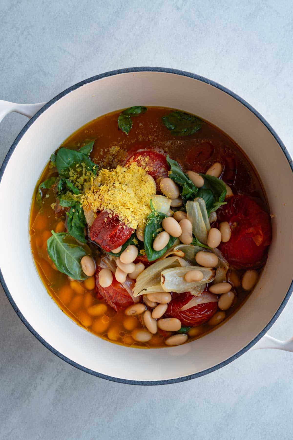 Roasted vine tomatoes, onion, garlic, as well as butter beans, nutritional yeast, and basil leaves in a large white saucepan.