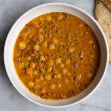 Orange colored chickpea and green lentil curry in a white bowl next to three pieces of naan.