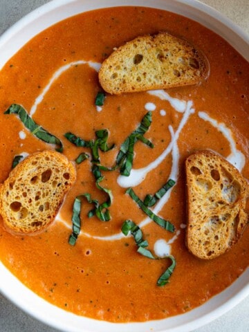 Creamy red tomato soup in a white bowl with croutons, chiffonade basil leaves, and a drizzle of vegan cream.