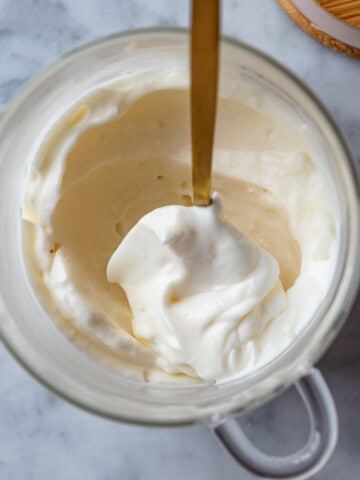 A glass jar with creamy white vegan mayonnaise inside being removed with a gold spoon, and a wooden lid beside it.