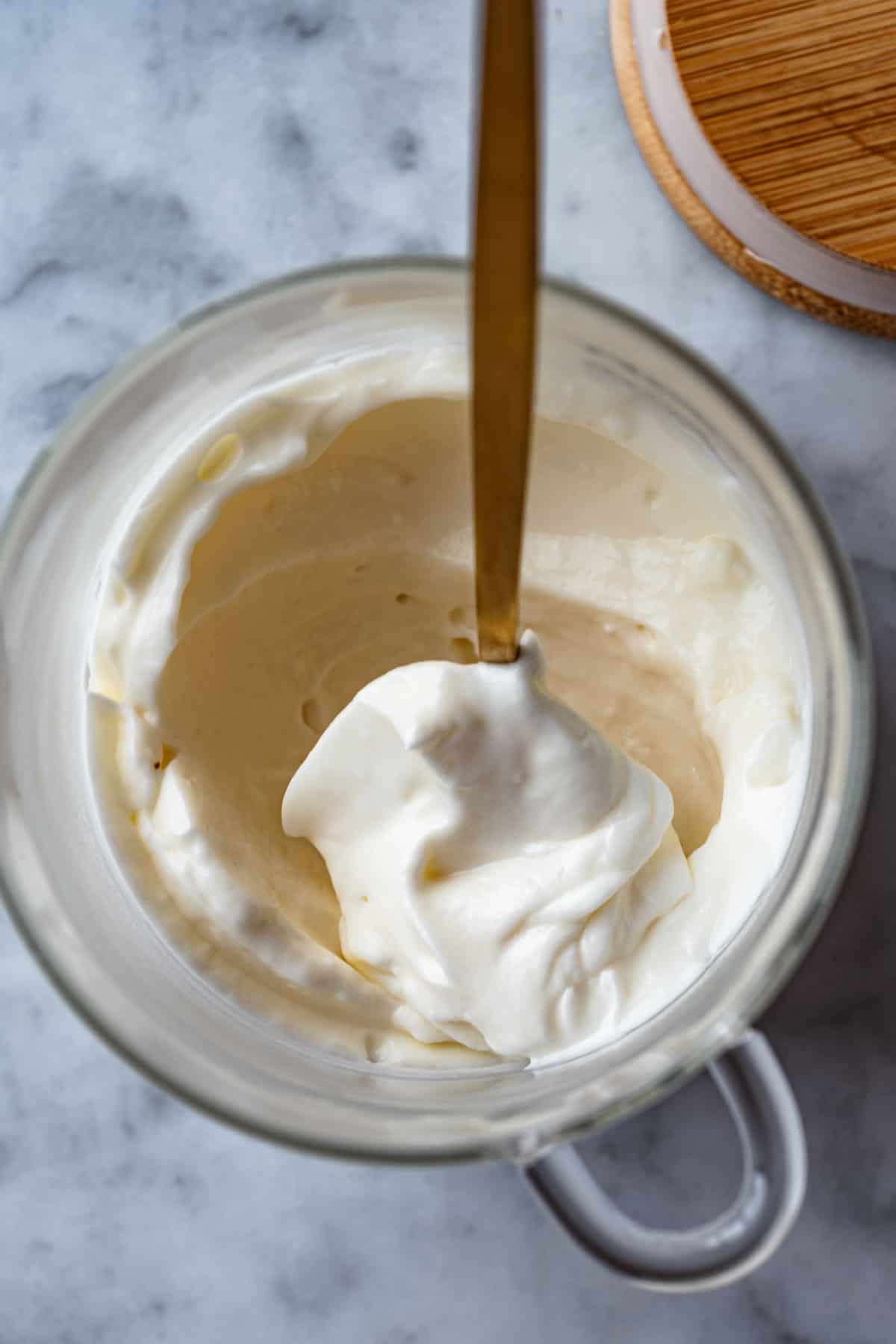 A glass jar with easy creamy white vegan mayonnaise inside being removed with a gold spoon, and a wooden lid beside it.