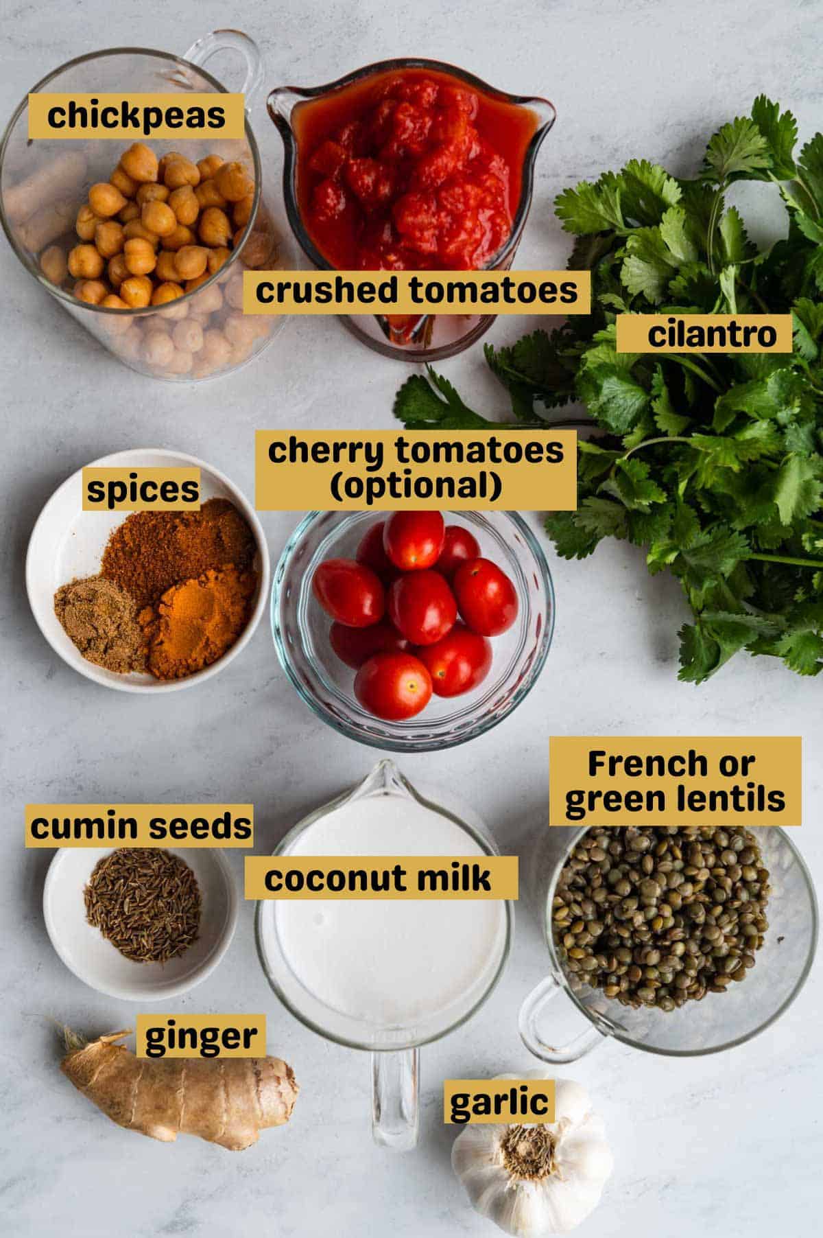 Chickpeas, crushed tomatoes, fresh cilantro, grape tomatoes, spices, cumin seeds, coconut milk, ginger root, garlic, and green lentils in glass containers on a white marble board.