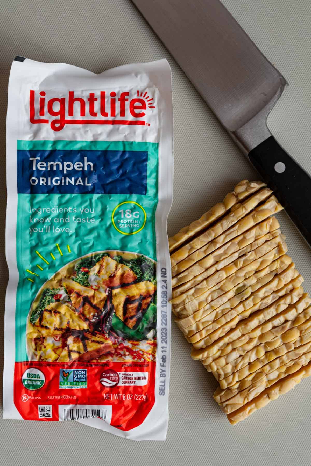 A packet of Lightlife tempeh with a block sliced thinly beside it and a chef's knife.