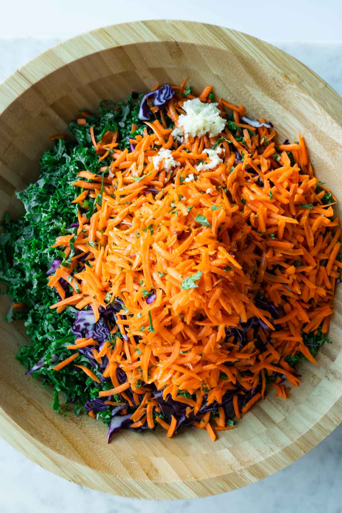 Sliced red cabbage, kale, shredded carrots, and finely pressed garlic in a large wooden bowl.