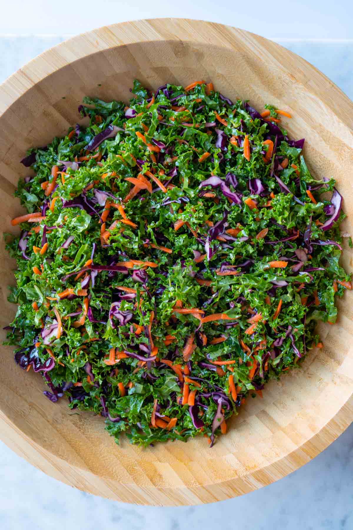 Sliced red cabbage, kale, and shredded carrots, tossed in oil dressing, in a large wooden bowl.
