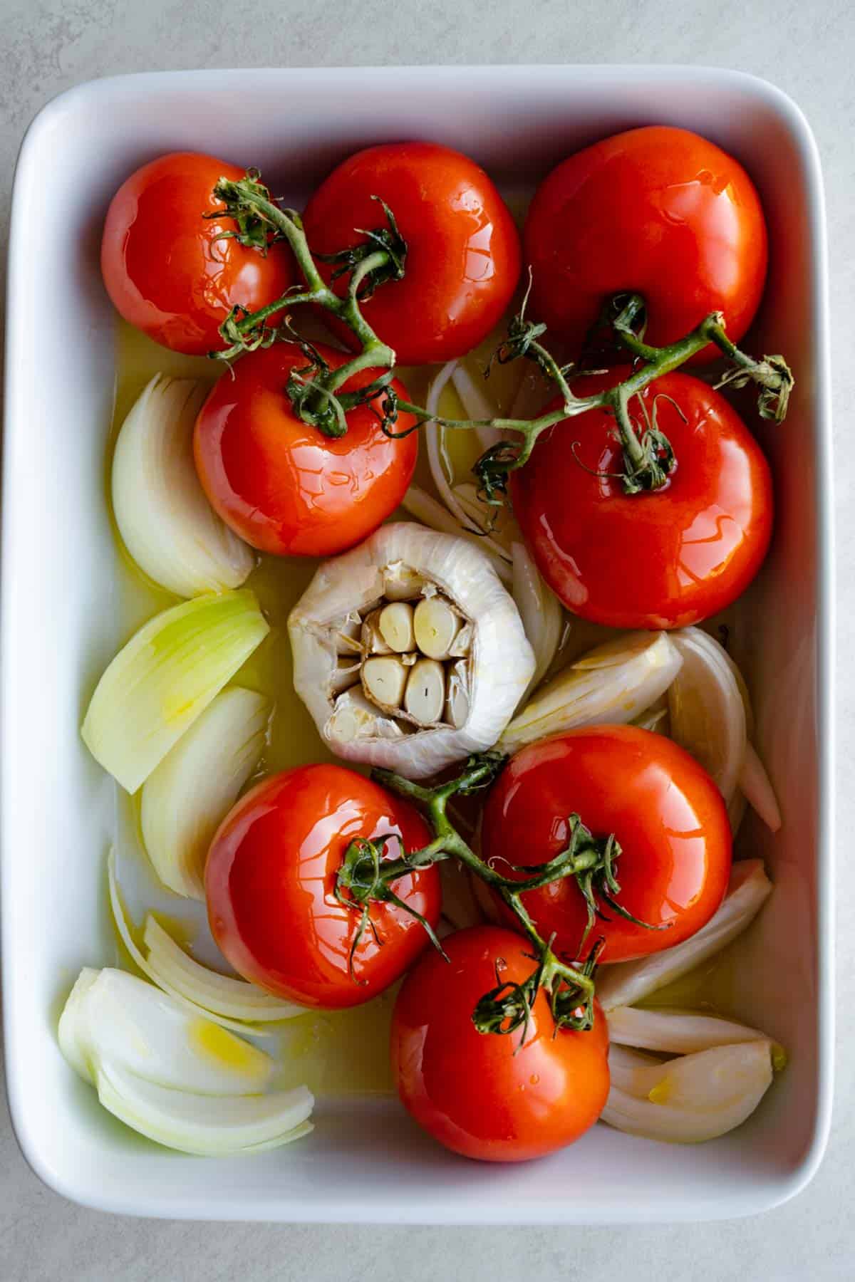 Eight vine tomatoes, chopped onion, and a head of garlic in a white baking dish with olive oil.