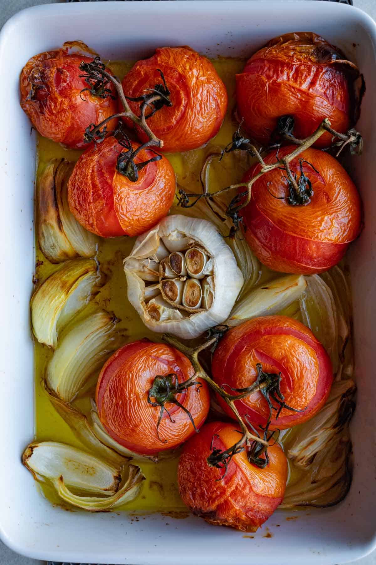 Roasted vine tomatoes, chopped onion, and a head of garlic in a white baking dish with olive oil.
