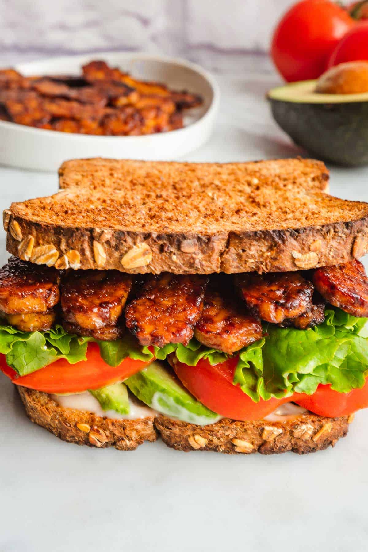 Vegan tempeh sandwich with tomato, lettuce, wholegrain bread, and avocado with a plate of tempeh bacon, one half avocado, and vine tomatoes in the background.