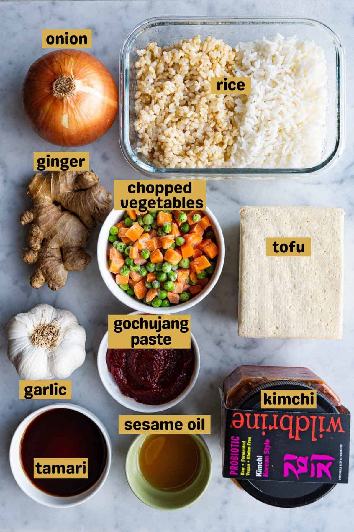One yellow onion, brown and white rice in a rectangular glass container, chopped frozen carrots and peas in a white bowl, ginger, one block tofu, garlic, gochujang paste, sesame oil, tamari, and kimchi on a marble board.