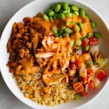 Quinoa, roasted onion, red bell pepper, and green bell pepper, baked tempeh, edamame, and kimchi drizzled with creamy pink chipotle dressing in a white bowl.