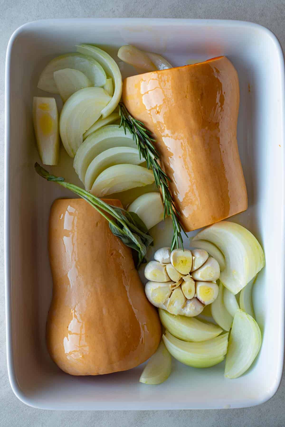 Halved butternut squash, flesh side down, with sliced onion, one head of garlic, and fresh sprigs of rosemary and sage in a white baking dish.