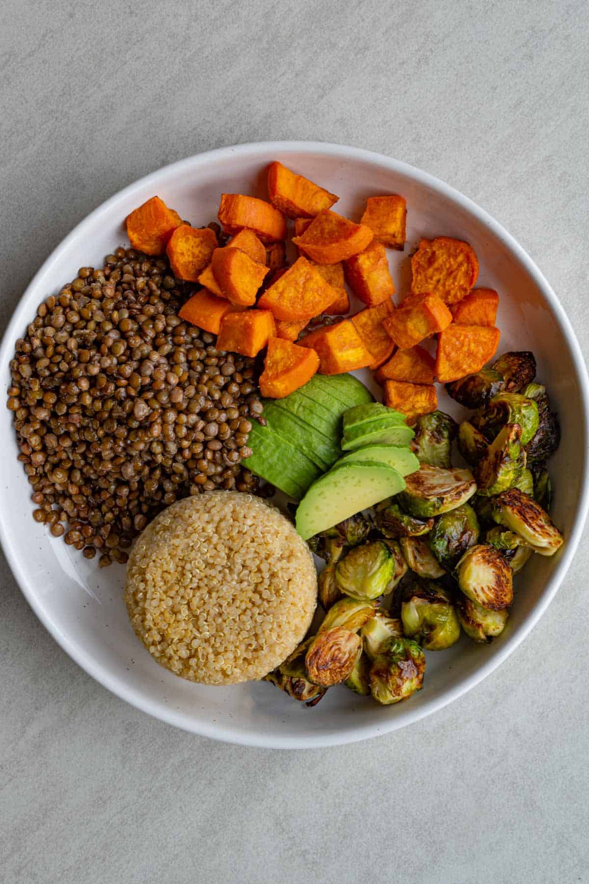 Roasted lentils, sweet potato, and Brussels sprouts, sliced avocado, and cooked quinoa in a white bowl.