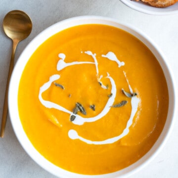 Orange creamy butternut squash soup in a white bowl with a drizzle of white vegan cream and a gold spoon and toasted bread.
