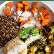 Roasted lentils, sweet potato, and Brussels sprouts, sliced avocado, and cooked quinoa drizzled with tahini dressing, and topped with hemp seeds in a white bowl.