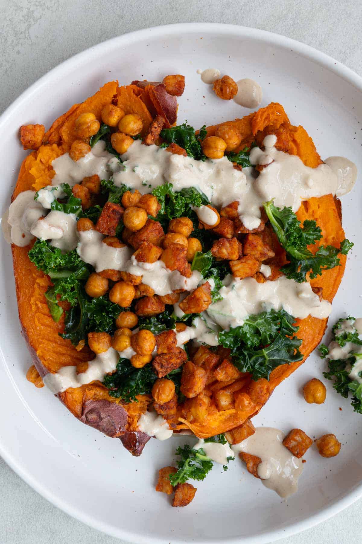 Loaded sweet potato with air fryer crispy tempeh and chickpeas, on a bed of massaged kale, with creamy tahini dressing in a white bowl.