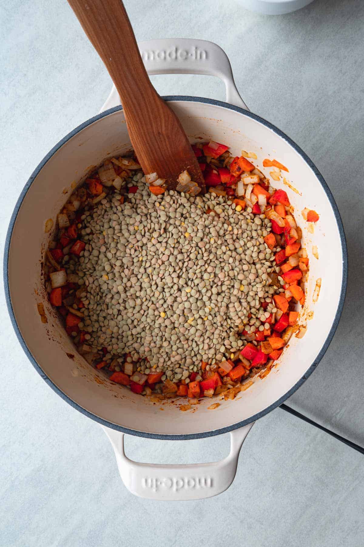 Chopped red bell pepper, carrots, onion, garlic, spices, and brown lentils cooking in a large white saucepan with a wooden spatula.