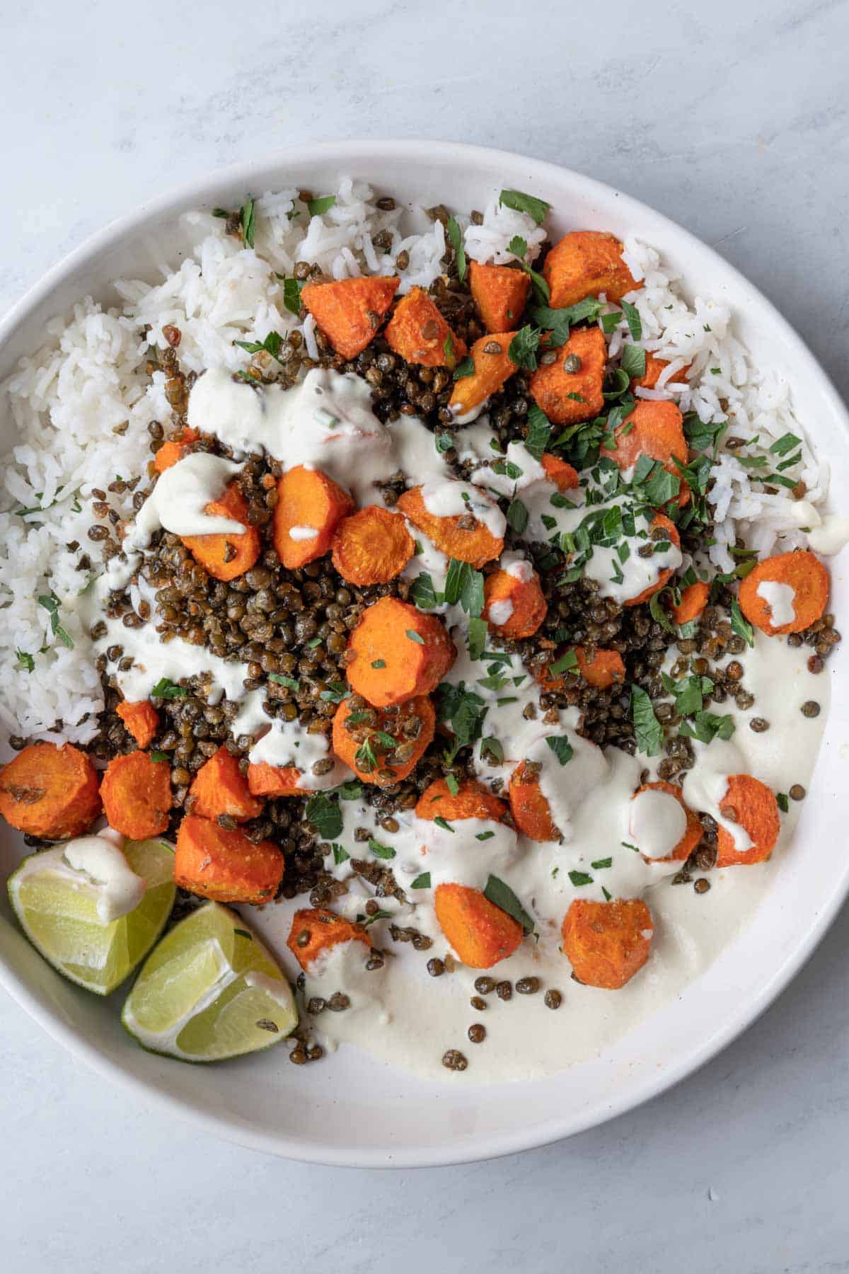 Buddha Bowl with roasted Puy lentils and carrot rounds on a bed of white rice with creamy white dressing, two lime wedges, and chopped parsley in a white bowl.