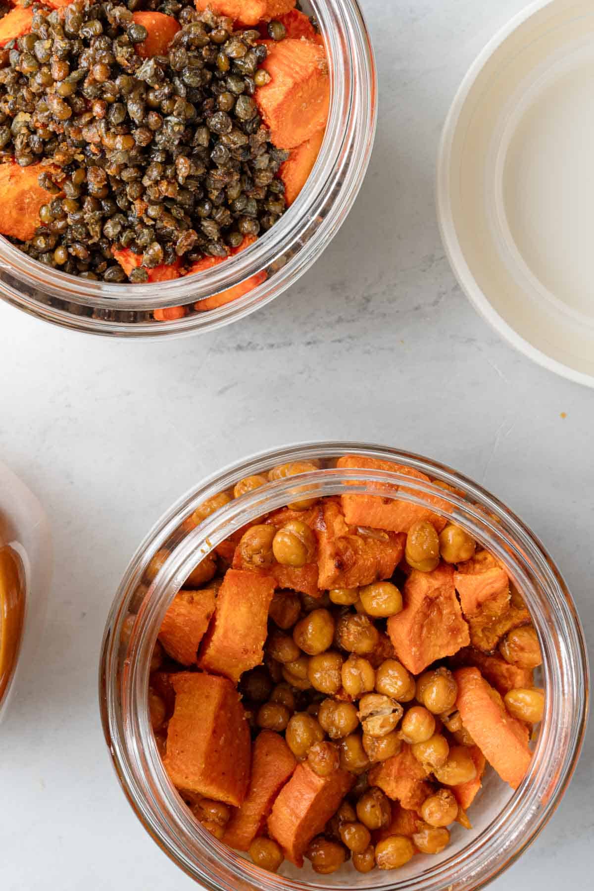 Two white round meal prep containers with Buddha Bowl meal prep. One with roasted chickpeas and sweet potatoes, and the other with roasted lentils and carrots.