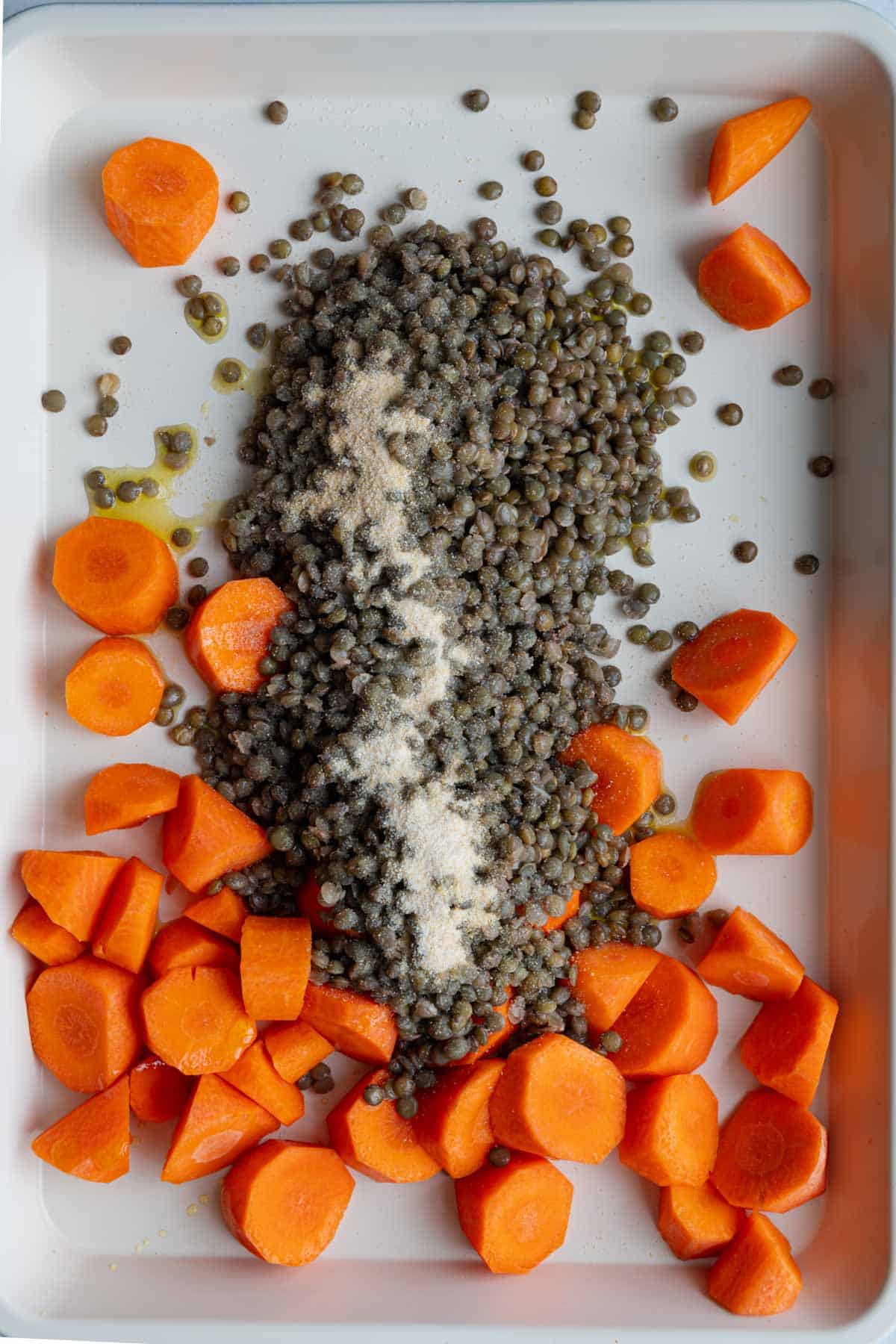 Puy lentils and cut carrot rounds on a white rimmed baking sheet with garlic powder and olive oil.