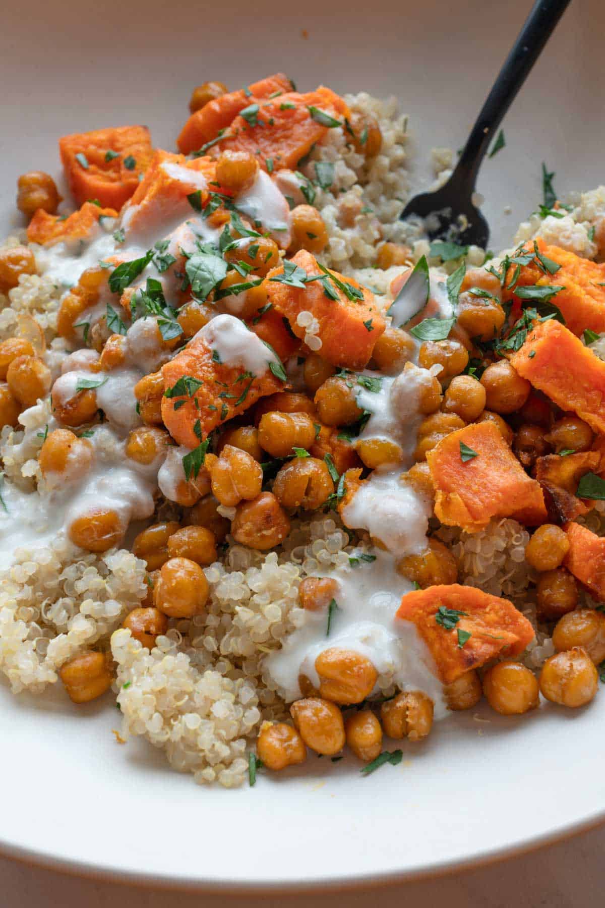 Sweet potato, roasted chickpeas, quinoa, white creamy garlic sauce, and a chopped parsley garnish with a black fork.