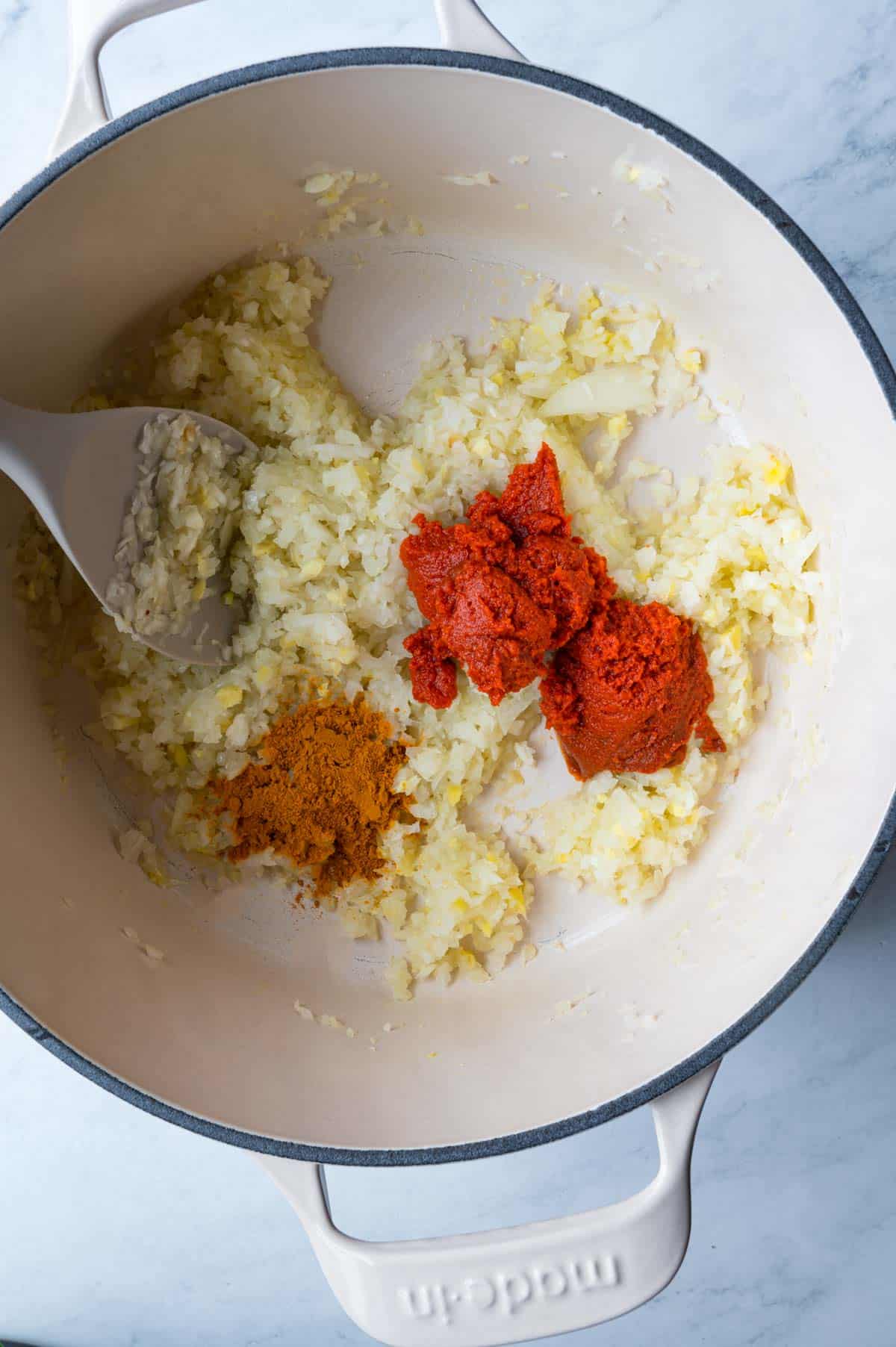 Cooked diced onions, minced garlic and ginger, with red paste and turmeric, in a white skillet with a gray spatula.