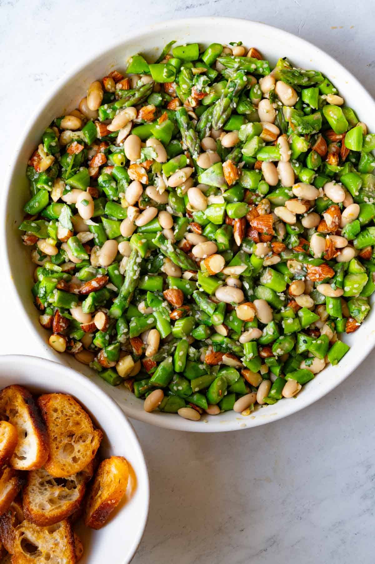 Chopped asparagus, white beans, almonds, miso dressing, and sesame seeds in a white bowl next to a small white bowl of crostinis.