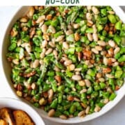 Chopped asparagus, white beans, almonds, miso dressing, and sesame seeds in a white bowl next to a small bowl with crostinis.