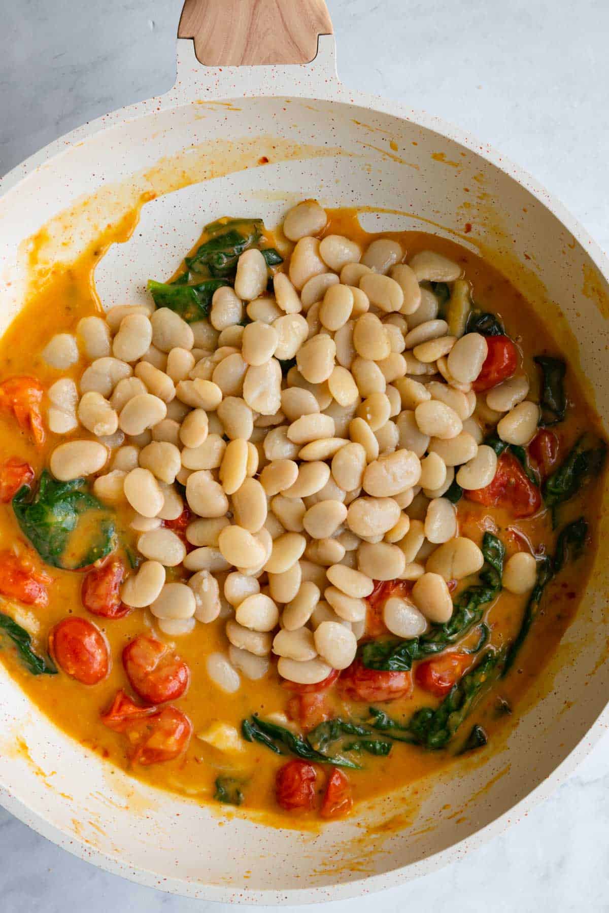 Vegan sauce in a white saucepan with burst grape tomatoes, and spinach with white beans on top.