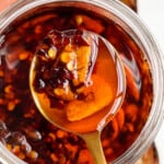 Chili crisp with crushed red pepper, garlic, shallots and oil on a gold spoon in a glass jar.