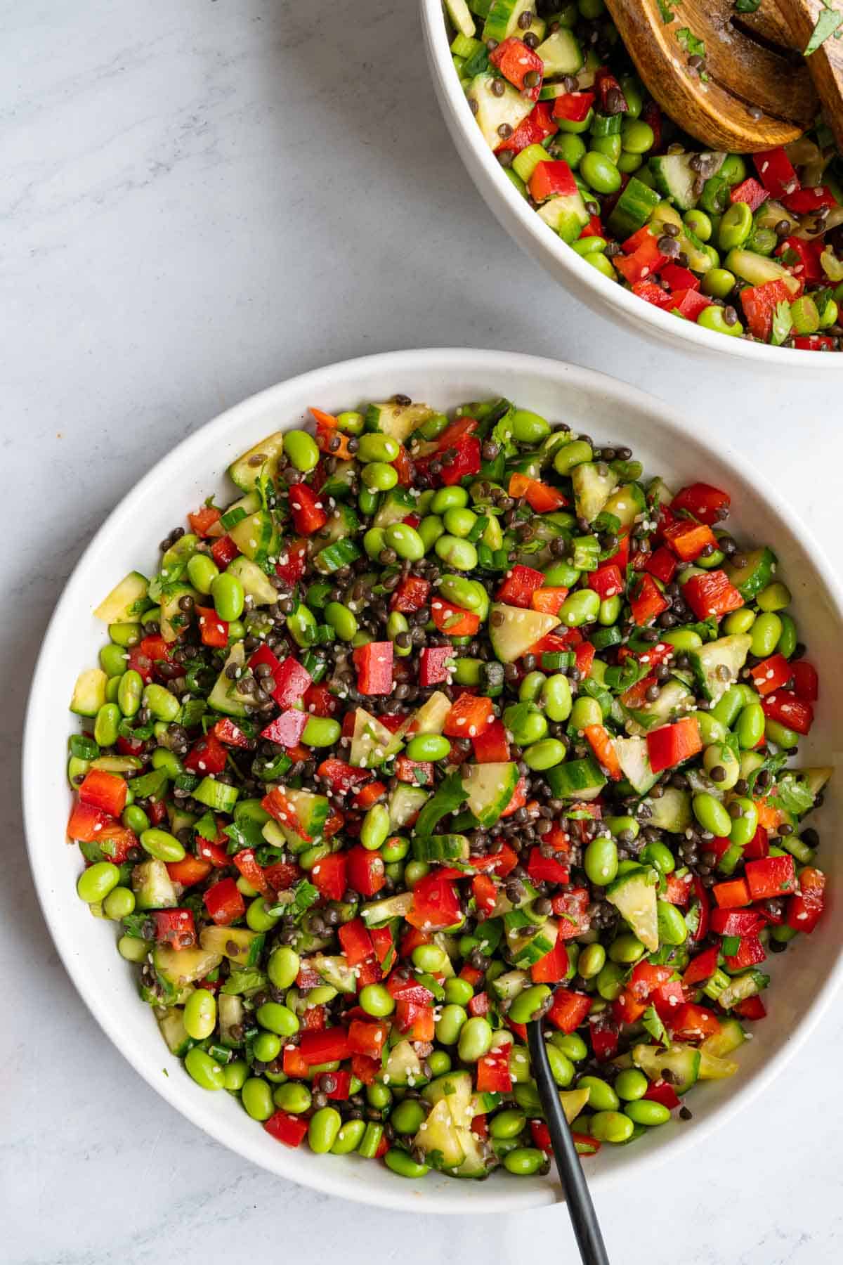Edamame, black lentils, chopped cucmber and red bell pepper, sesame seeds, and chopped green onion and cilantro, in a white bowl with a black fork, next to another bowl with more edamame salad and wooden servers.