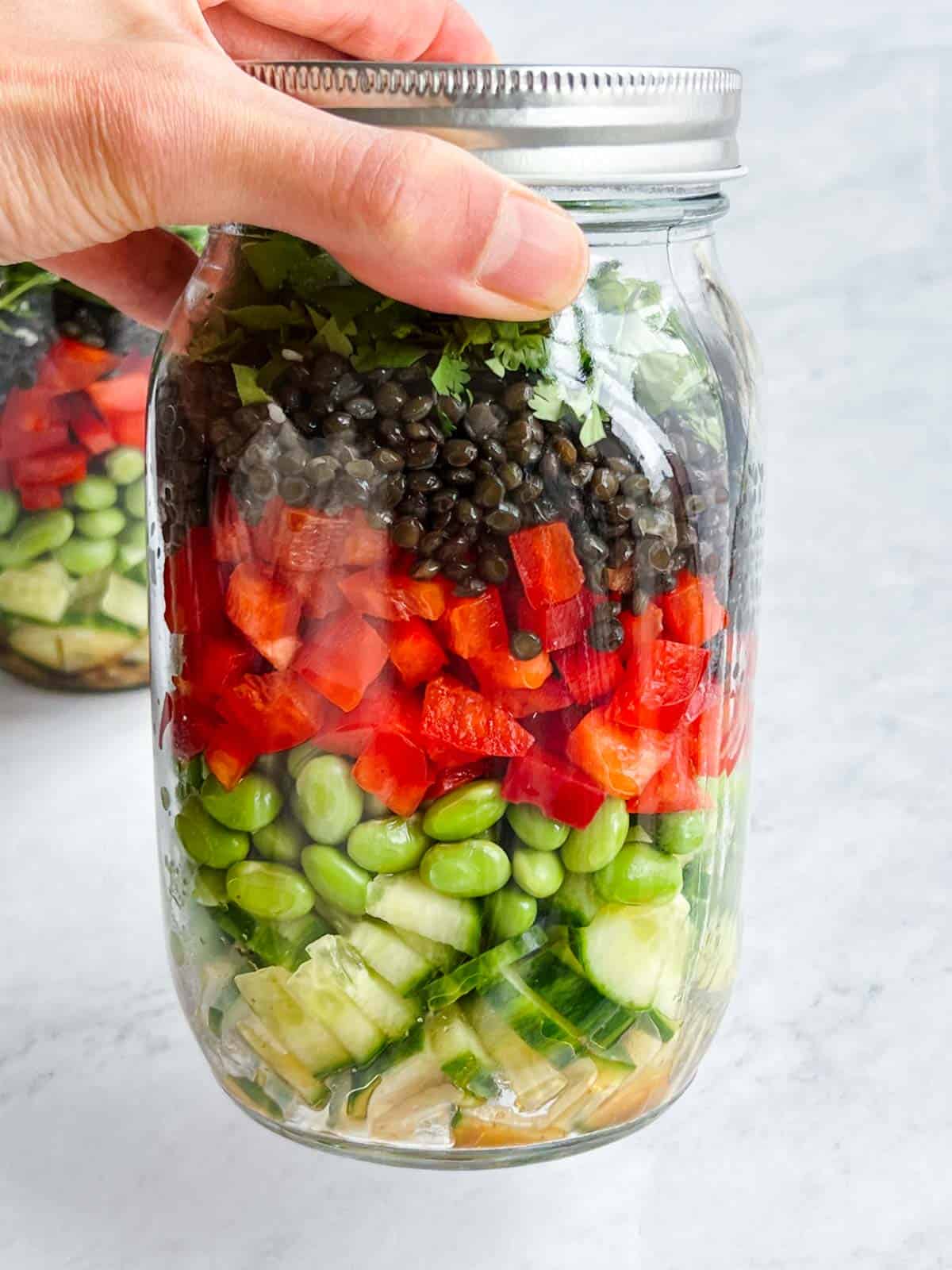 A layered mason jar with dressing at the bottom, then chopped cucumber, edamame, chopped red bell pepper, black lentils, chopped cilantro, and sliced green onion, with another filled jar in the background.