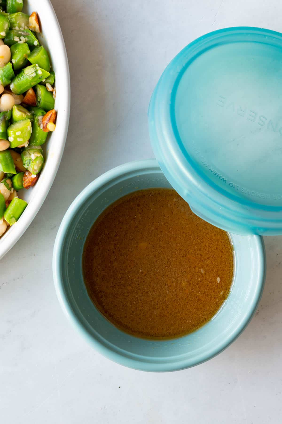 Miso dressing in a small turquoise meal prep container next to a white bowl with chopped veggies.