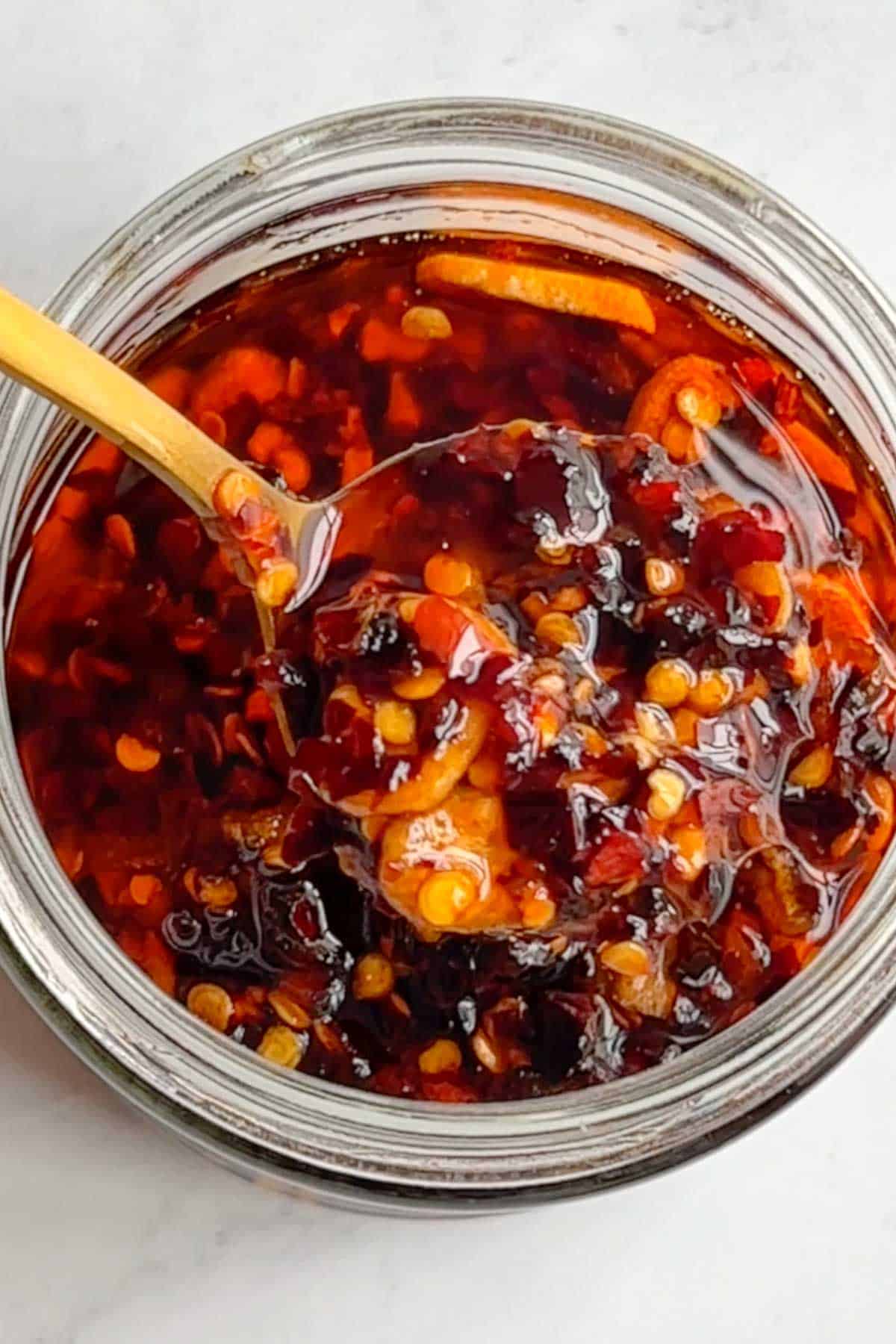 Chili crisp with crushed red pepper, garlic, shallots and oil on a gold spoon in a glass jar.
