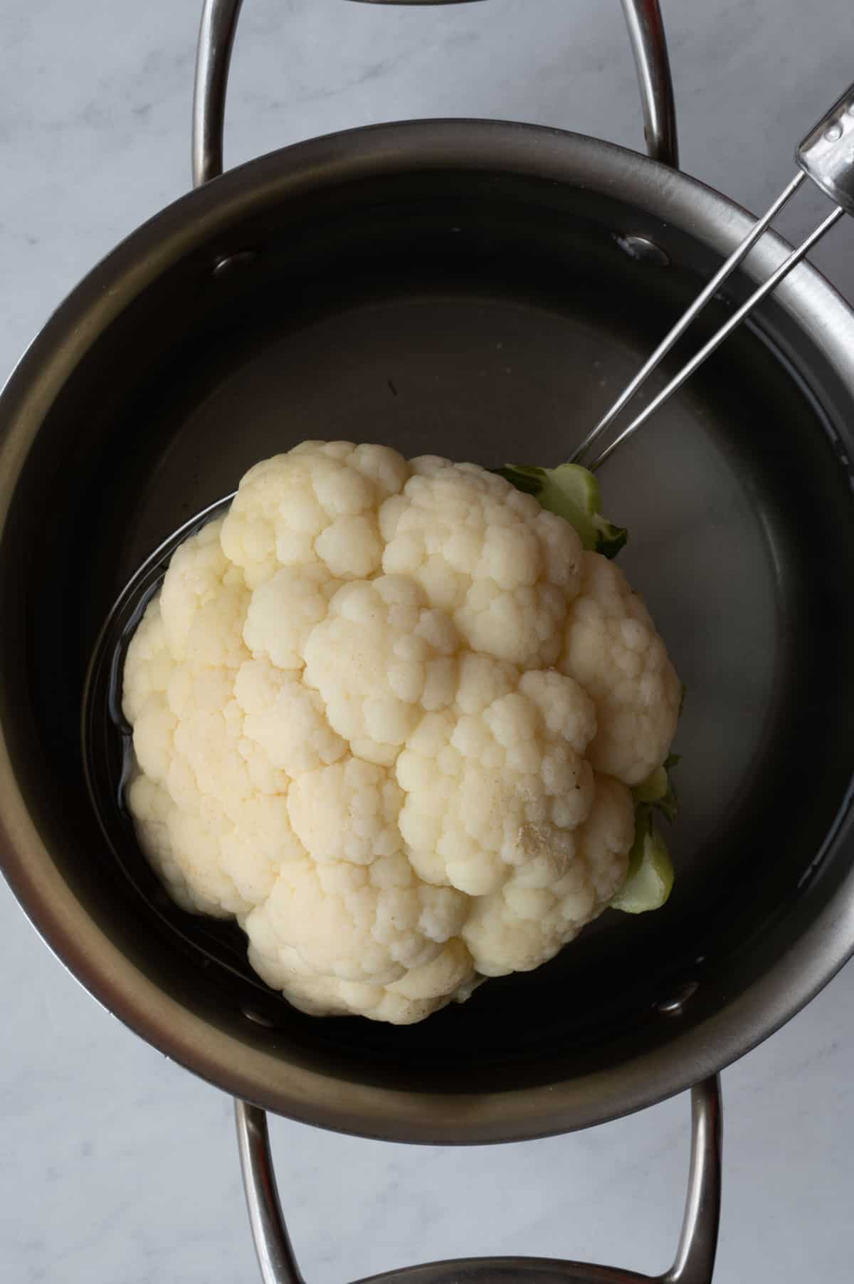 One medium head cauliflower in a spider coming out of a stainless steel saucepan with water.