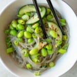 Easy noodle salad with sesame dressing, edamame, cucumber, crushed red pepper, and green onion in a white bowl and black chopsticks.