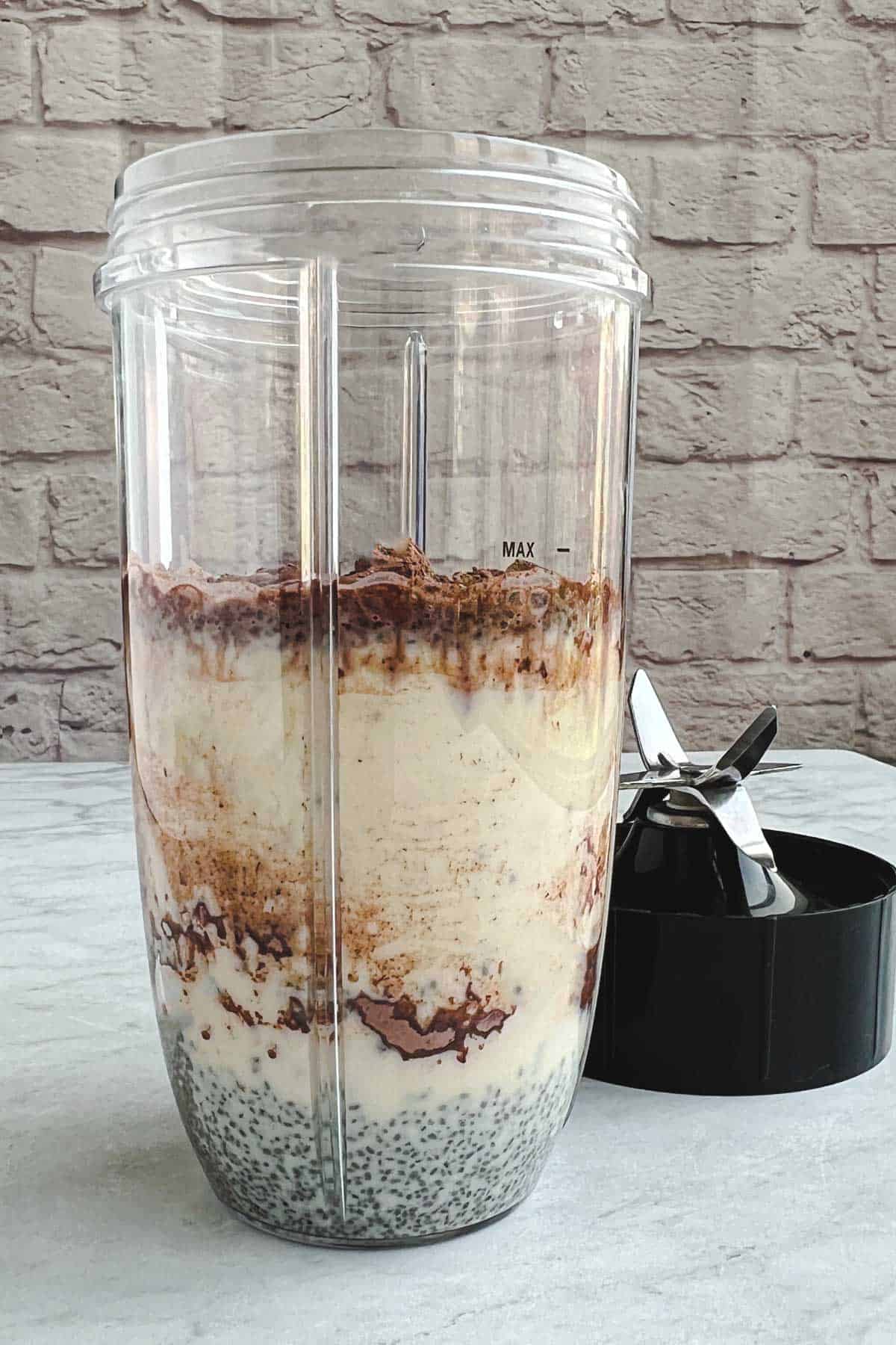 A plastic blender canister with chia seeds, almond milk, cocoa powder, dates, and vanilla with the black lid with blade on the right.