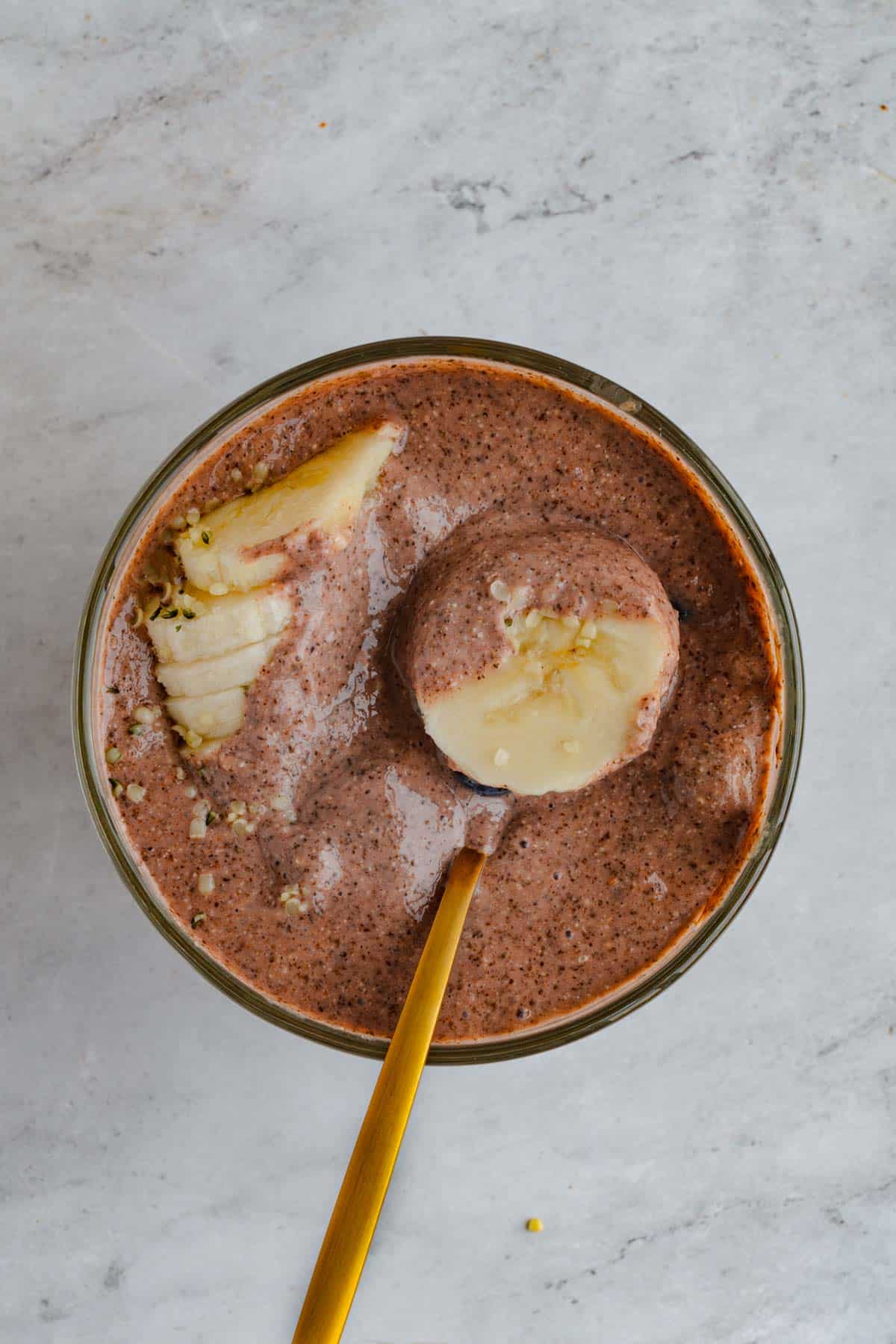 Blended chocolate chia pudding in a glass jar topped with banana slices with a gold spoon inside.