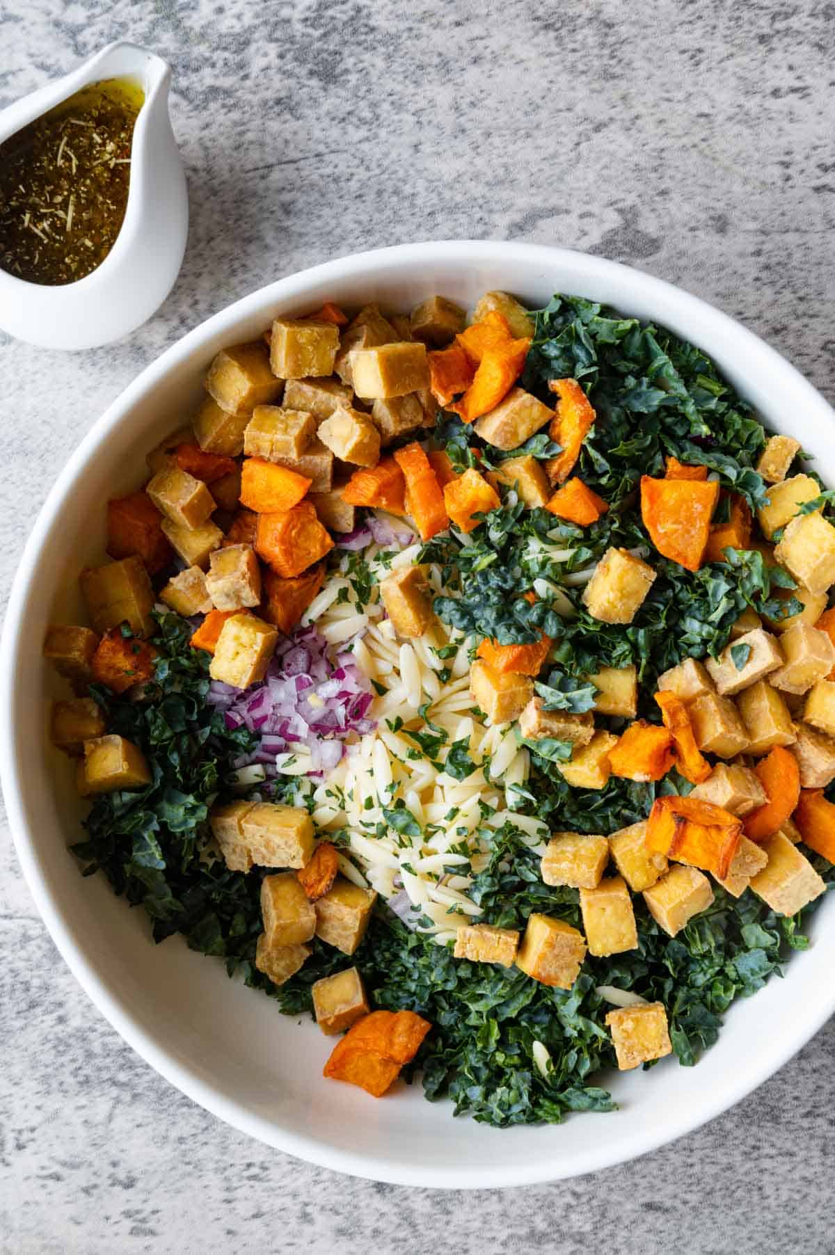 Orzo, crispy tofu, chopped kale, roasted sweet potato, chopped red onion, and roasted pepitas in a white bowl with a white pouring bowl of dressing on the side.