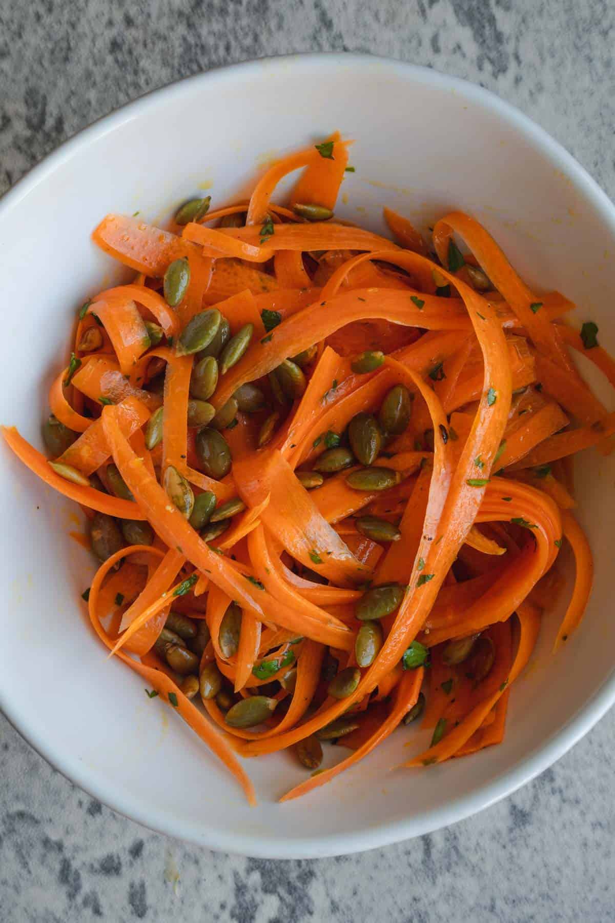 Raw carrot salad in a white bowl with roasted pepitas, chopped herbs and dressing.