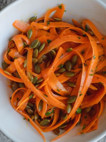 Raw carrot salad in a white bowl with roasted pepitas, and chopped herbs.