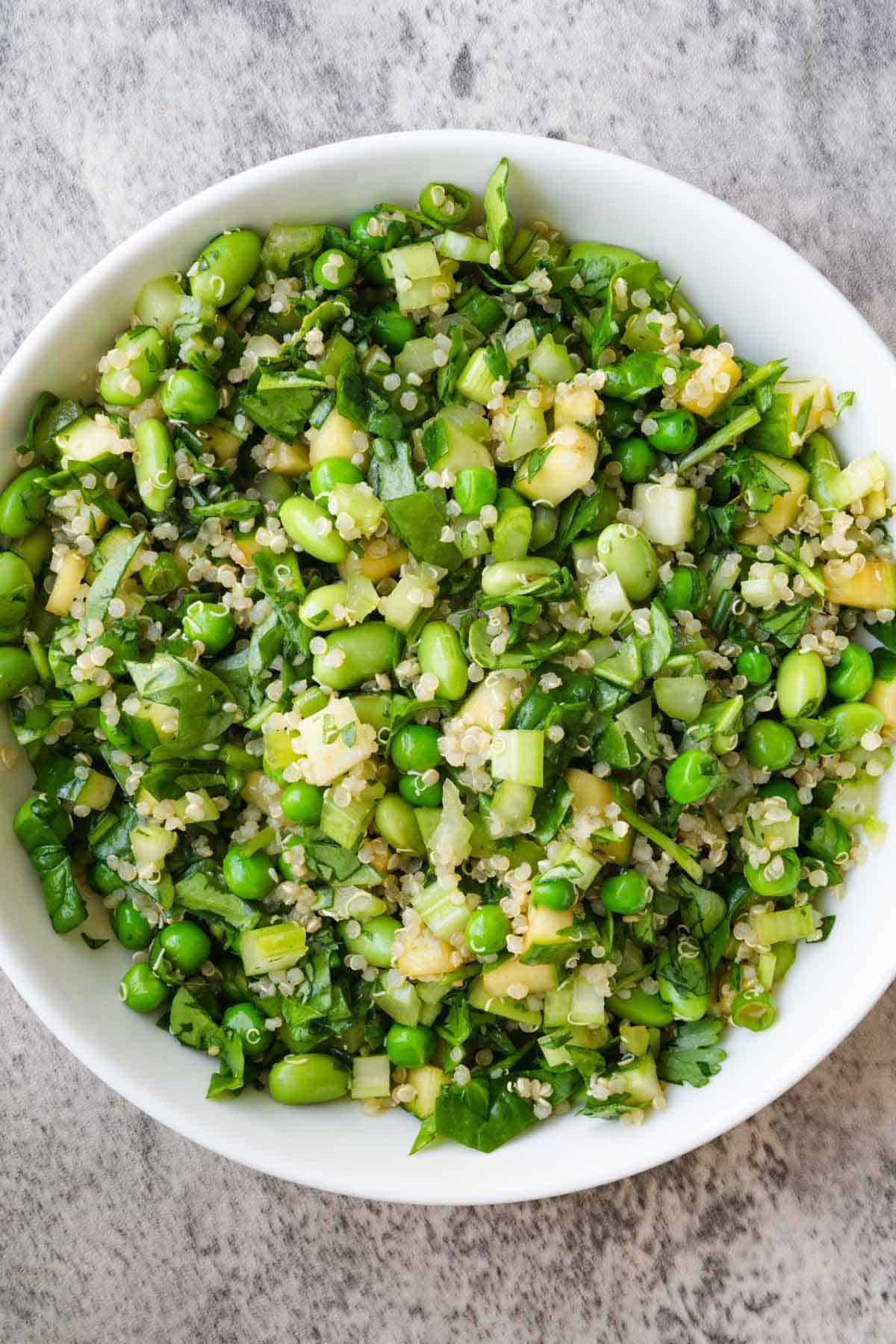 A white bowl of salad with Asian dressing, cucumber, celery, green peas, edamame, quinoa, chopped spinach and cilantro.