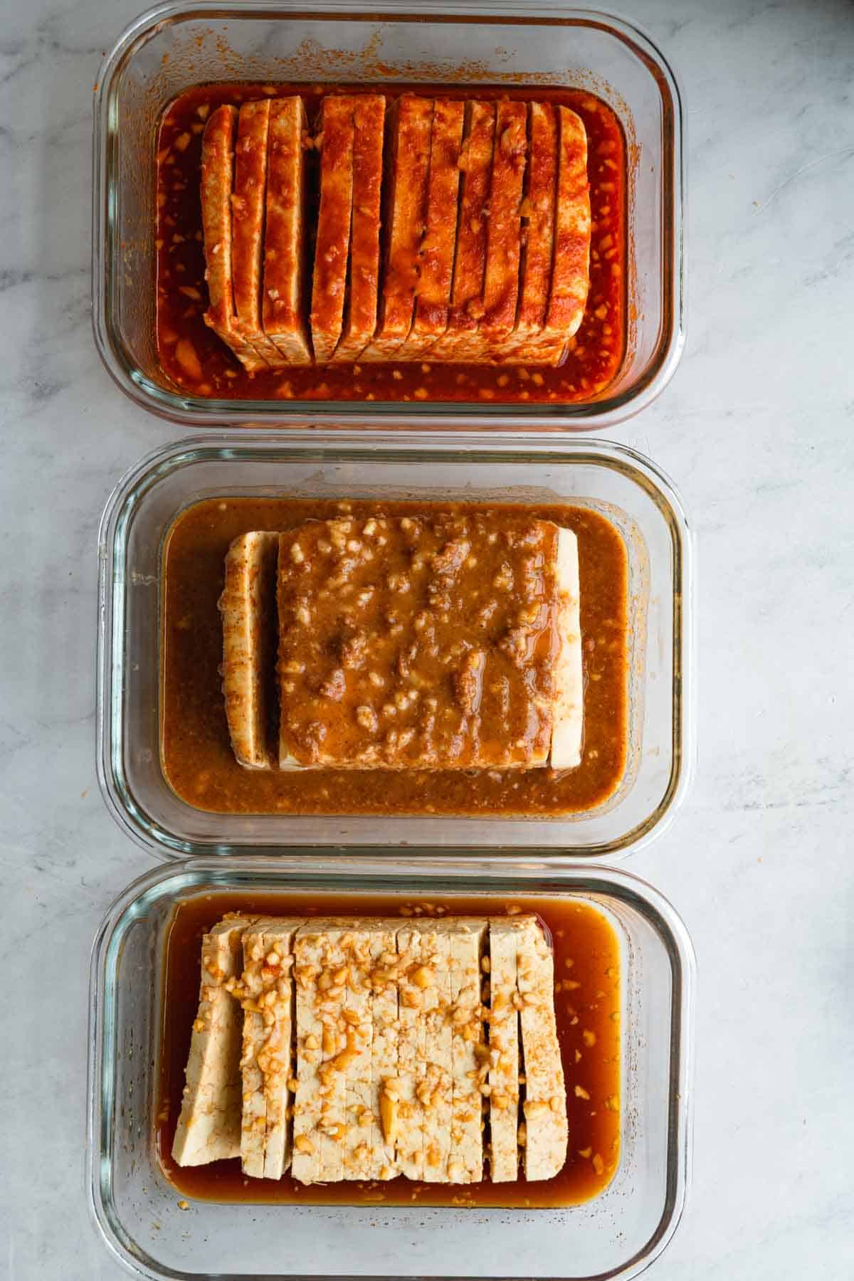 Three blocks of sliced tofu with three different marinades in three separate glass containers.