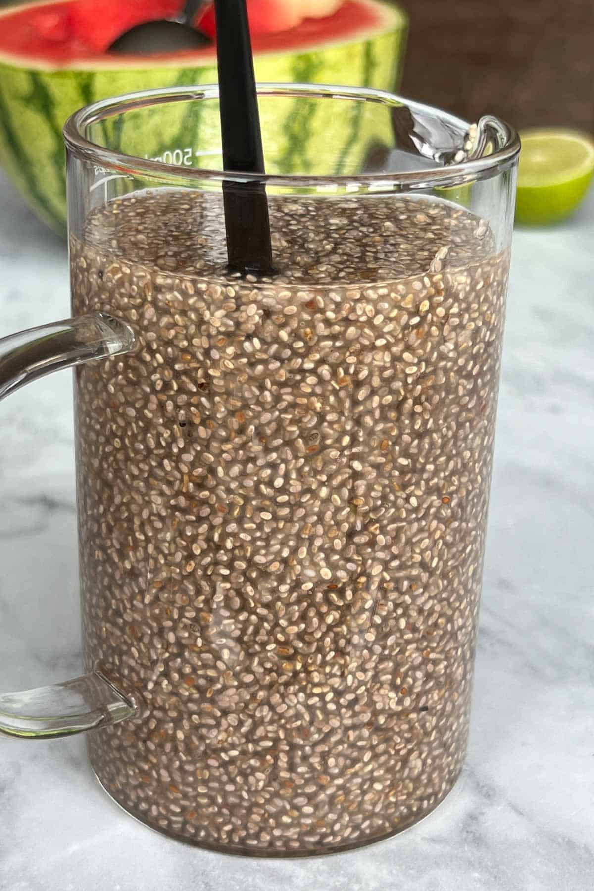 Chia seeds in a measuring jug with water gelling with a black spoon. Plus, one lime and one half watermelon in the background.
