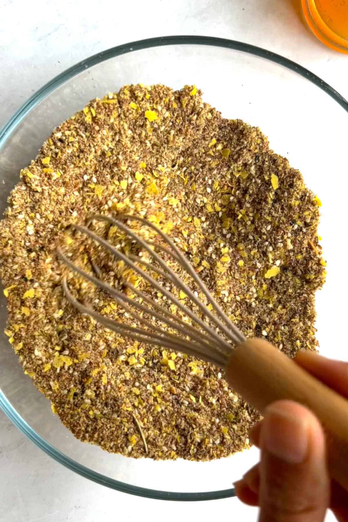 Nutritional yeast, ground flax seeds. sesame seeds and seasoning in a glass bowl with a grey silicone whisk.