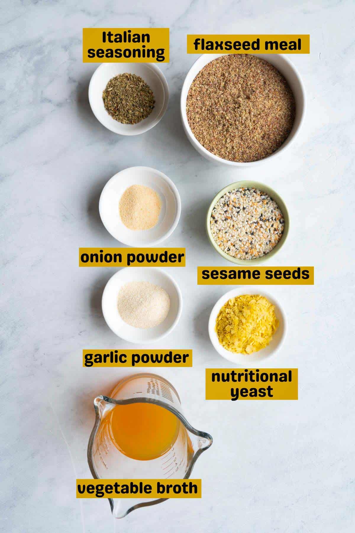 Italian seasoning, flaxseed meal, onion powder, sesame seeds, garlic powder, nutritional yeast, and vegetable broth in containers on a white backdrop.