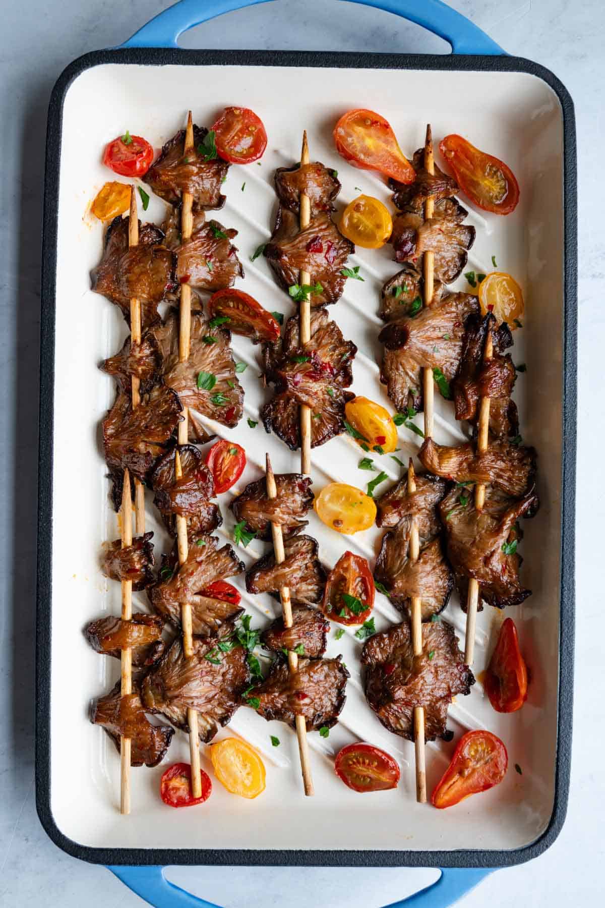 Nine oyster mushroom skewers on a white and blue grilling dish with grape tomatoes and chopped parsley.